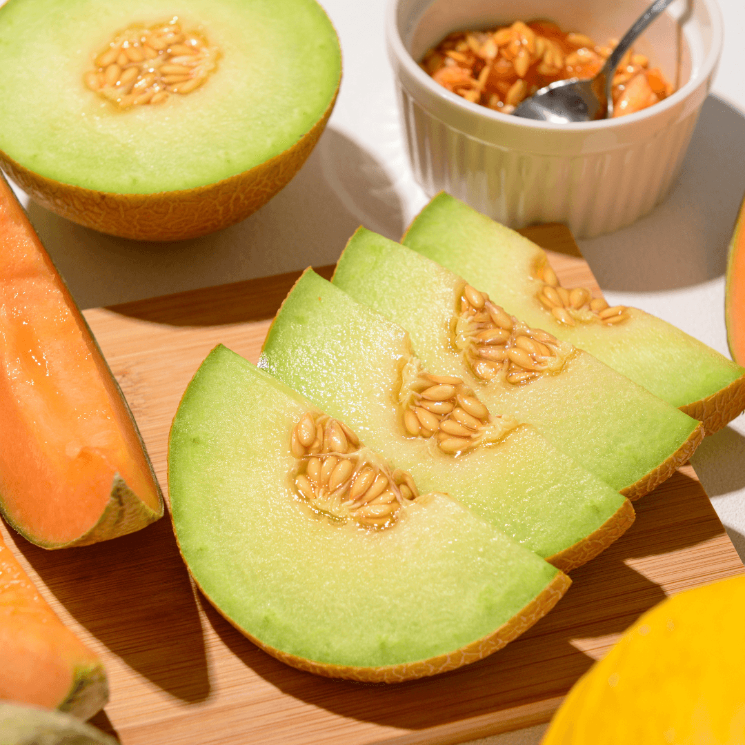 Sliced green honeydew melon displaying orange seeds, arranged on a wooden board with whole melons and a white bowl in the background, lit by natural sunlight.