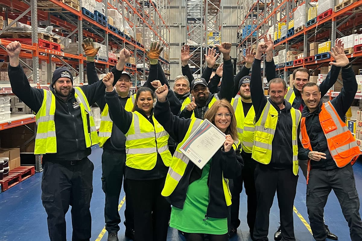 A group of warehouse workers in high-visibility vests celebrating indoors, with one person holding a framed BRC certificate, surrounded by industrial racking and storage shelves.