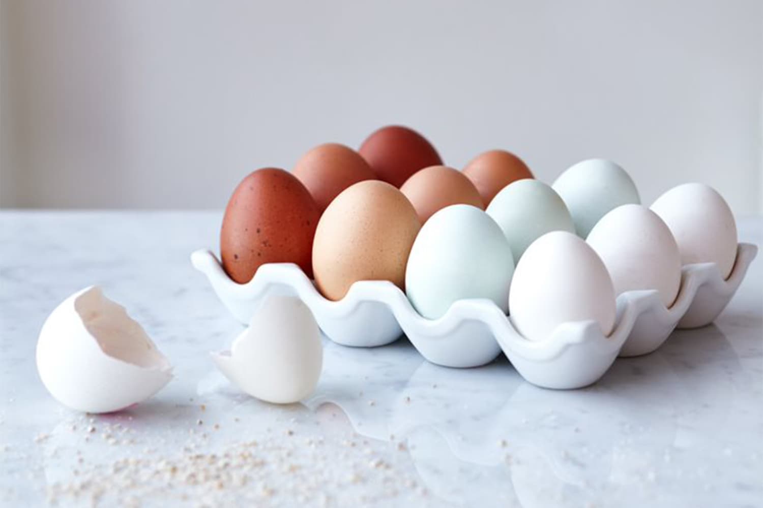 A collection of fresh eggs in various shades including dark brown, light brown, pale blue and white eggs, nestled in white wavy ceramic egg cups, with one broken eggshell in the foreground on a marble surface.