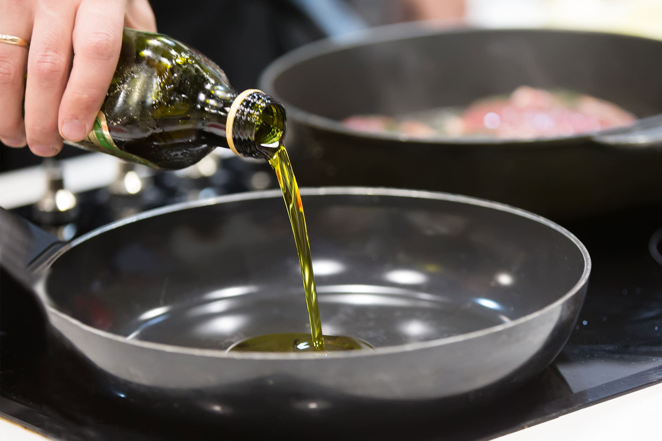 A person pouring golden olive oil from a dark glass bottle into a shallow pan on a stovetop, with another pan visible in the background containing food being cooked.