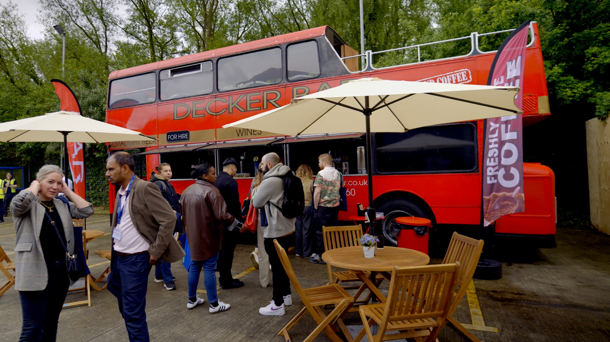 A red double-decker bus converted into a mobile food and beverage service, with customers gathered around serving counters and outdoor seating under umbrellas.