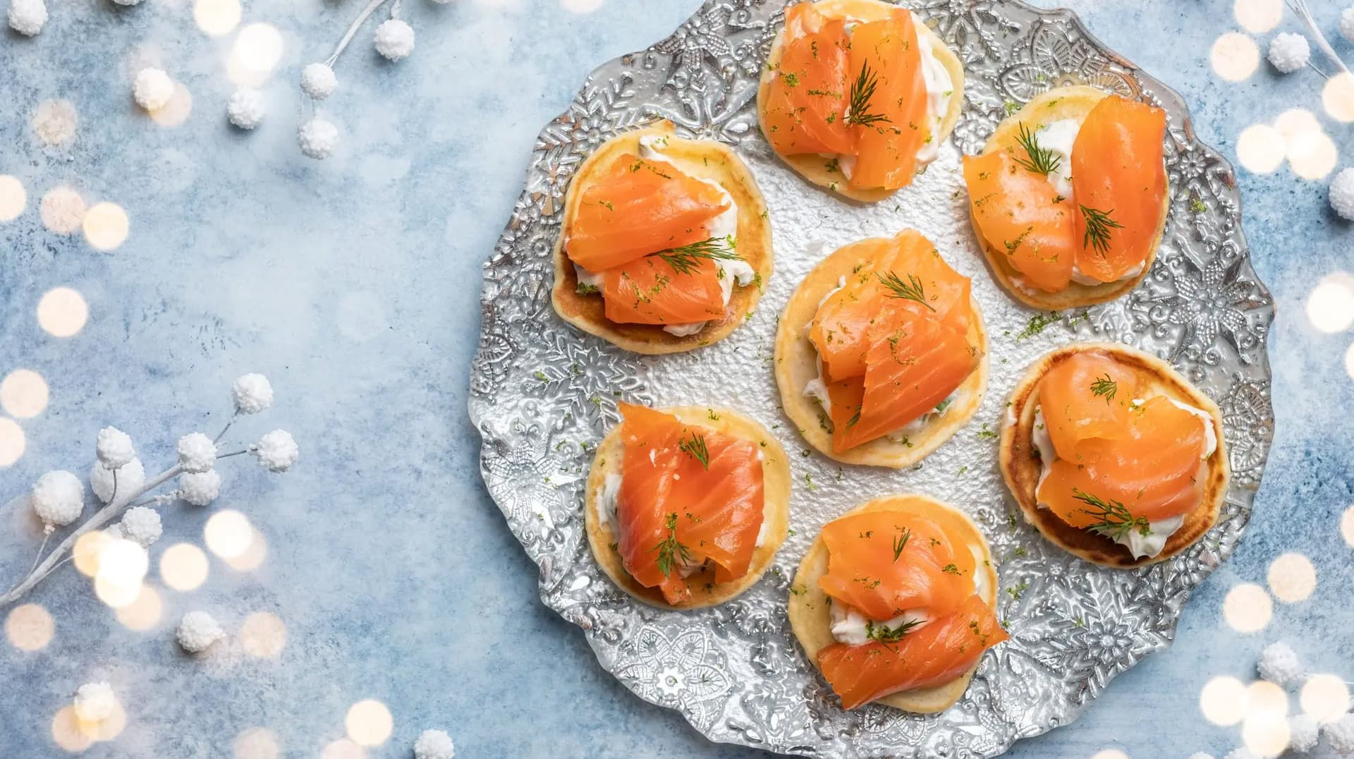 Seven elegant smoked salmon canapés arranged on an ornate silver plate, each topped with cream cheese and fresh dill, displayed on a blue textured surface with white decorative berries.
