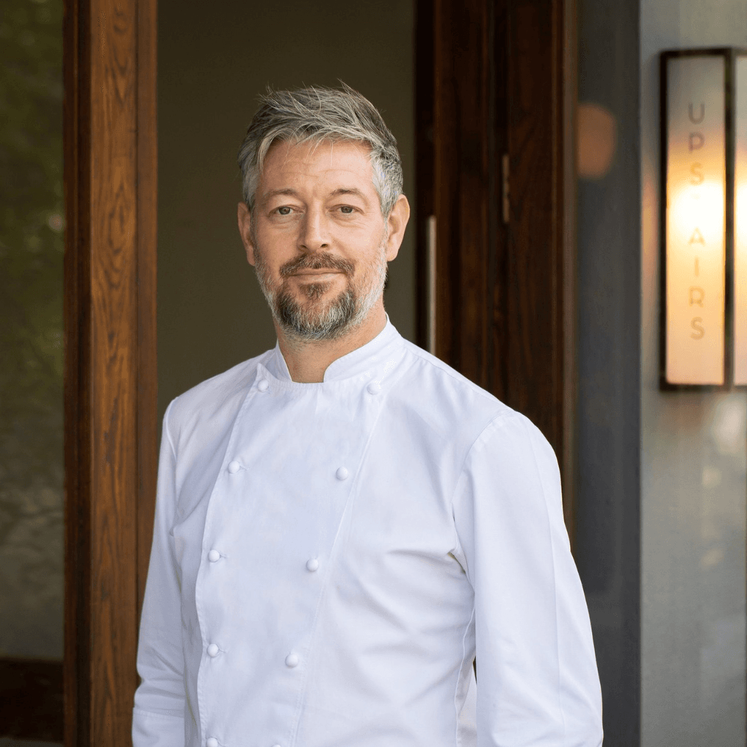 A middle-aged man with grey hair and beard wearing a white double-breasted chef's coat, photographed in a modern restaurant corridor with wooden panelling and framed artwork.
