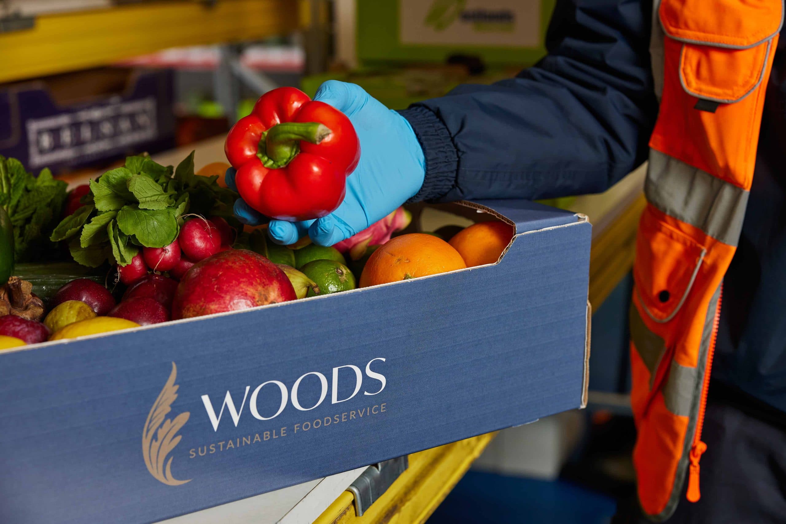 A worker in blue gloves holding a red bell pepper above an open Woods Sustainable Foodservice box filled with fresh fruits and vegetables including apples, oranges, radishes, and herbs.