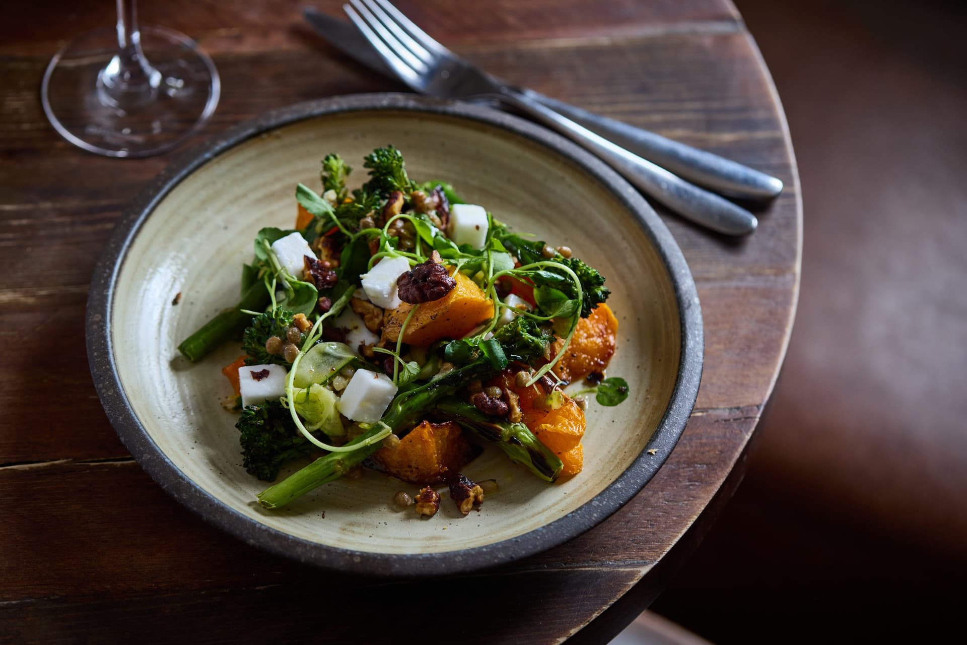A beautifully plated salad featuring roasted butternut squash, fresh greens, white cheese cubes, and garnishes served in a ceramic bowl on a wooden table with cutlery and glassware.