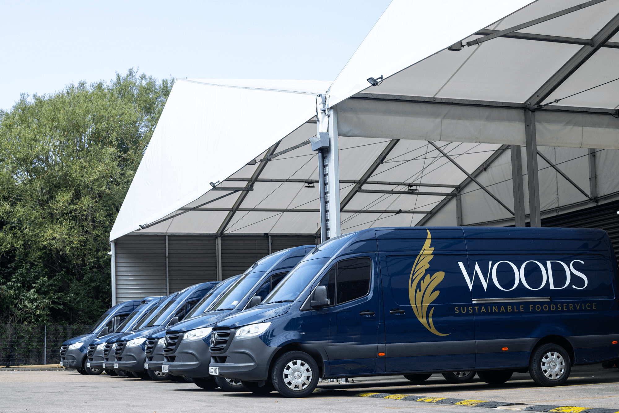 A row of six blue and grey branded delivery vans from Woods Sustainable Foodservice parked in a line, with large warehouse structures visible in the background.