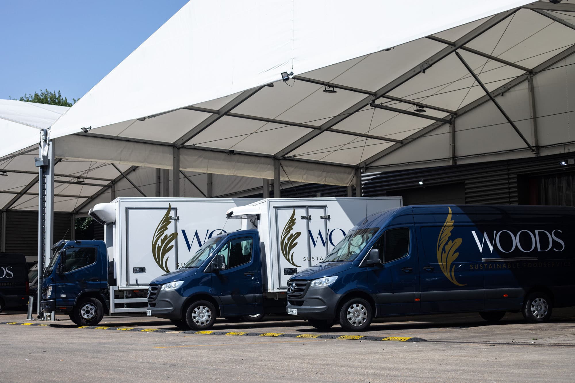 Three blue and white branded delivery vans parked under a large industrial canopy structure at a Woods Foodservice facility.