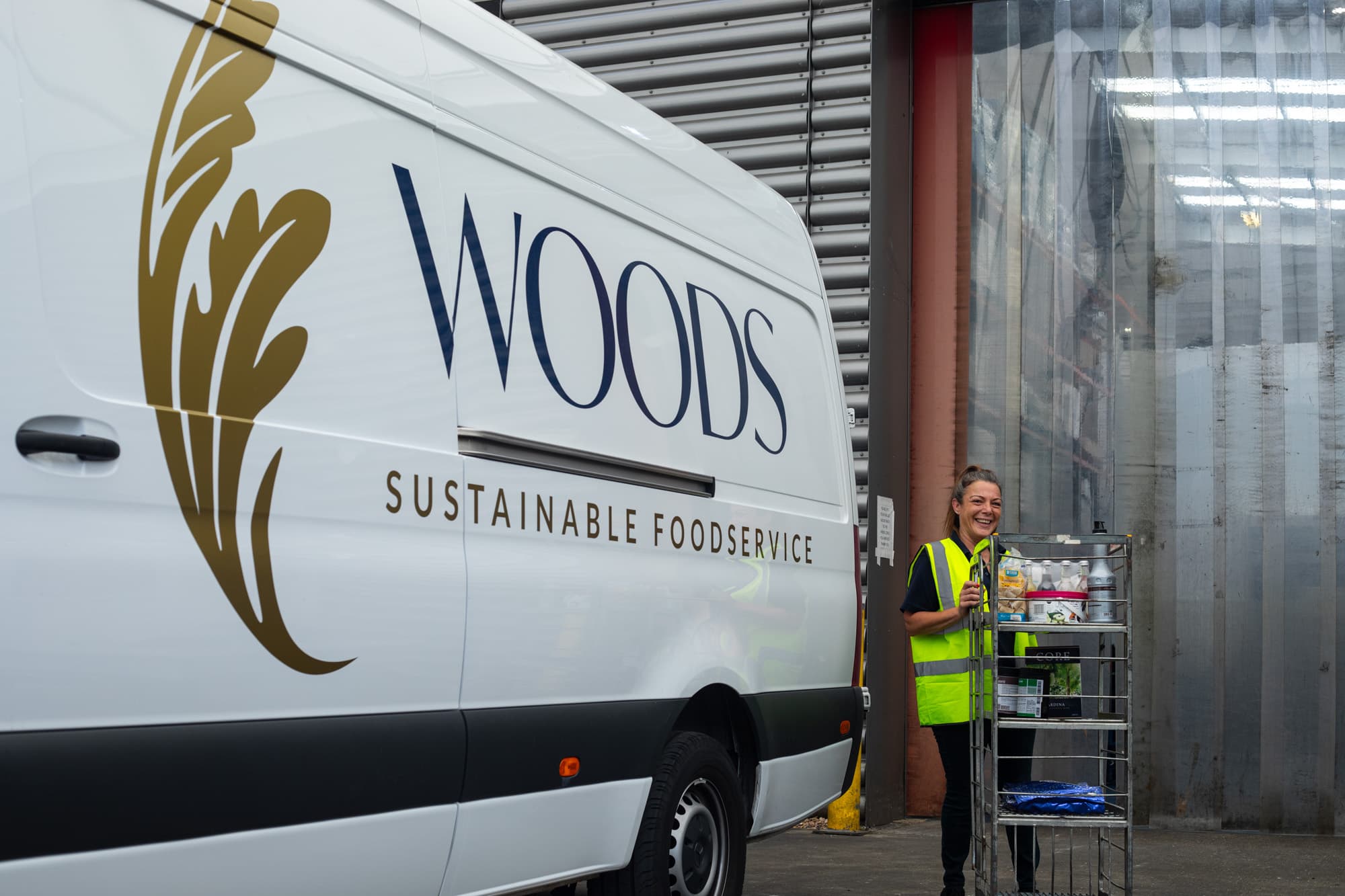 A female staff member in a high-visibility yellow vest standing next to a white Woods Sustainable Foodservice delivery van, loading or unloading food items from a metal shelving unit.