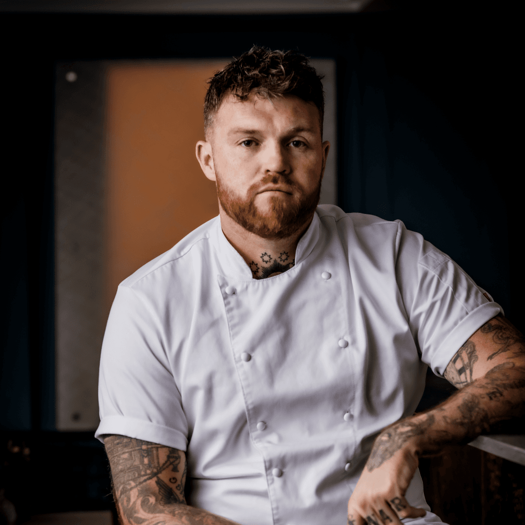 A bearded man with tattoos wearing a white short-sleeved chef's coat, photographed against a blurred warm-toned background in a professional studio setting.