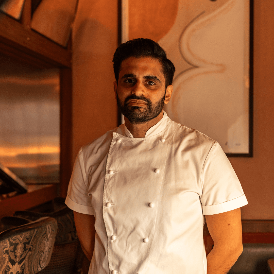 A bearded man wearing a white short-sleeved chef's coat, photographed indoors against a warm-toned orange and cream background.