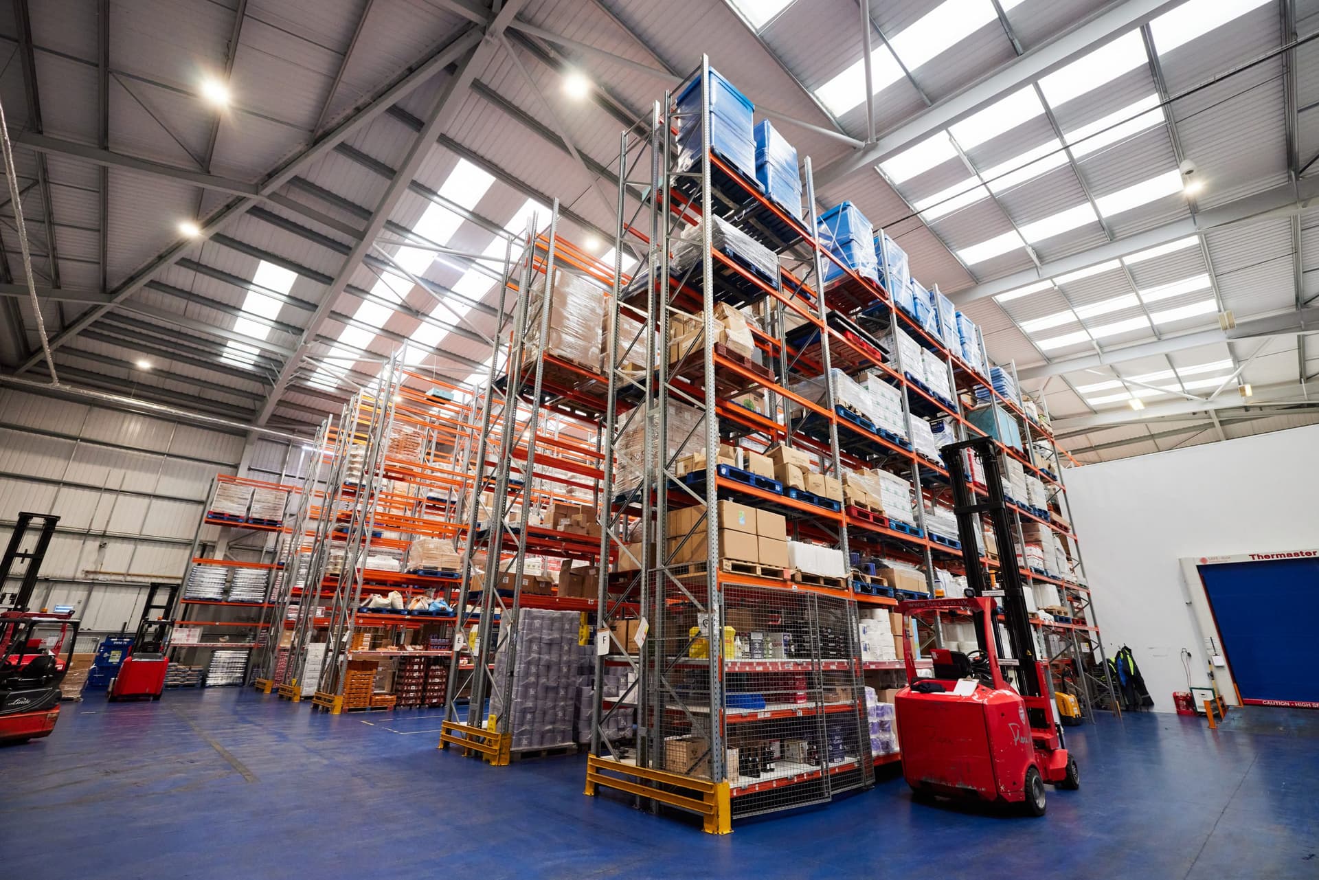 Interior of a large warehouse with high pallet racking systems storing blue and white boxes, red forklift visible, industrial ceiling with skylights.