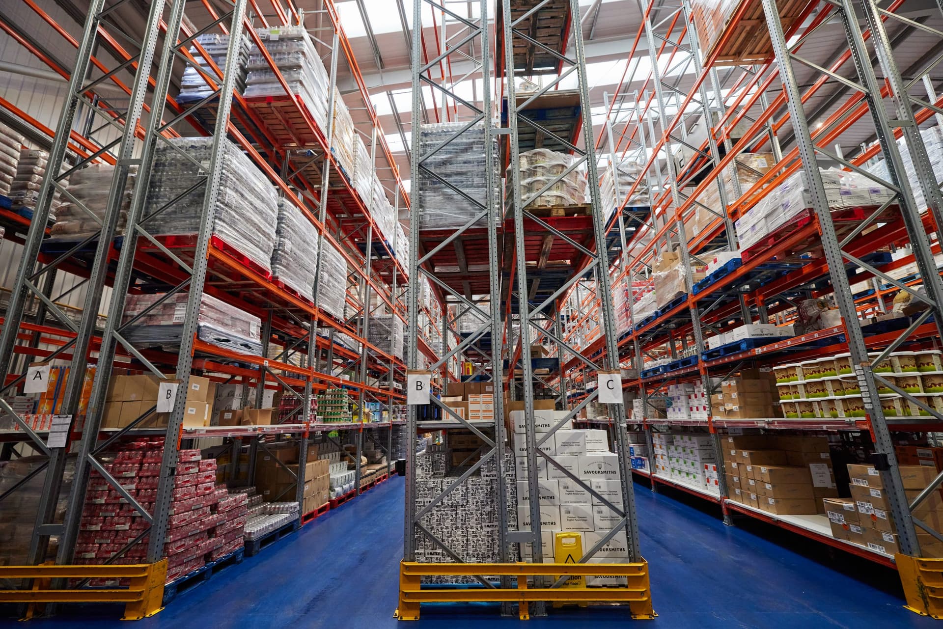 Interior view of a large food storage warehouse featuring multiple levels of industrial metal racking with red and orange beams, stacked cardboard boxes and packaged products, blue flooring, and safety signage.