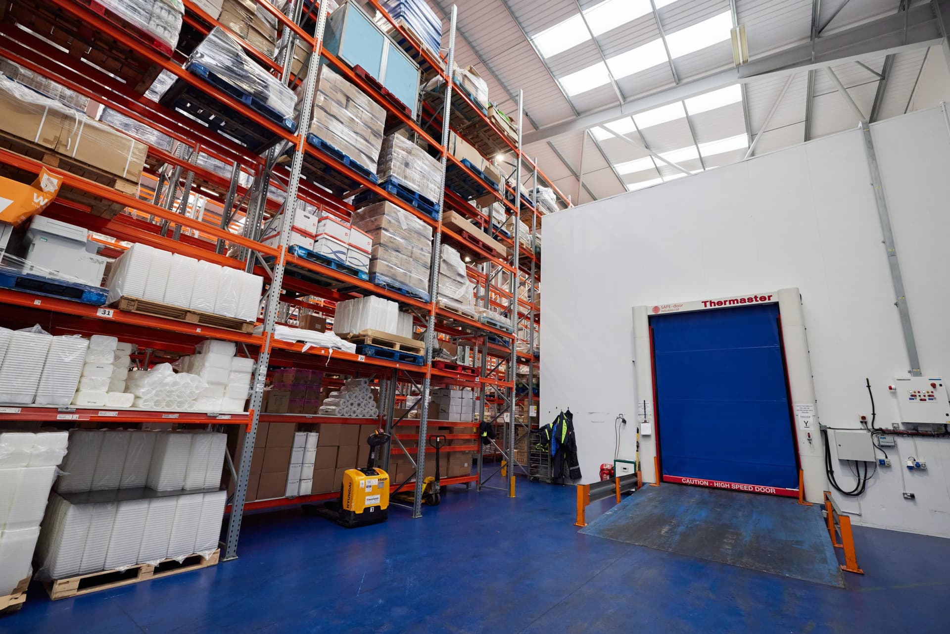 Industrial warehouse facility interior showing orange pallet racking systems stocked with packaged goods, blue epoxy flooring, skylight ceiling, and a white wall with blue high-speed door labelled Thermaster.
