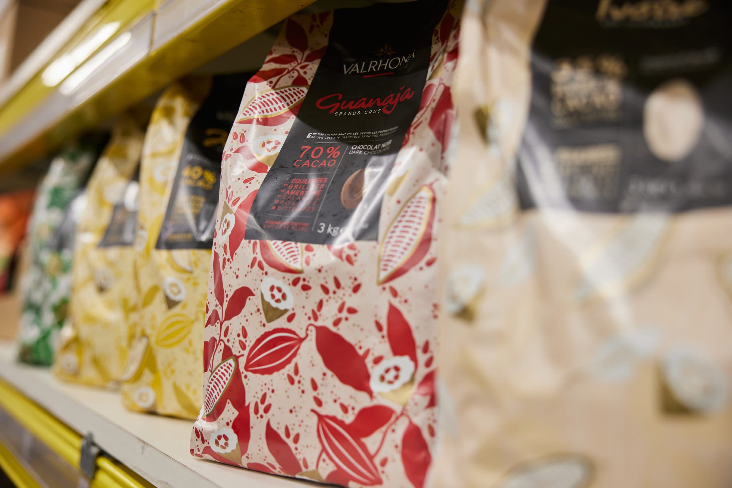 Valrhona Guanaja dark chocolate package displayed on a yellow retail shelf alongside other chocolate products, showing red and cream patterned packaging with cocoa pod design.