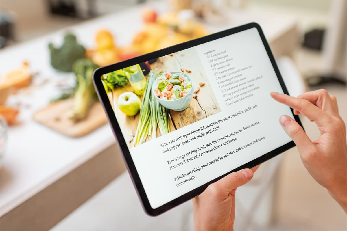 A person holding a tablet displaying a recipe with ingredient list and instructional photos, positioned over a kitchen counter with fresh vegetables and prepared ingredients.