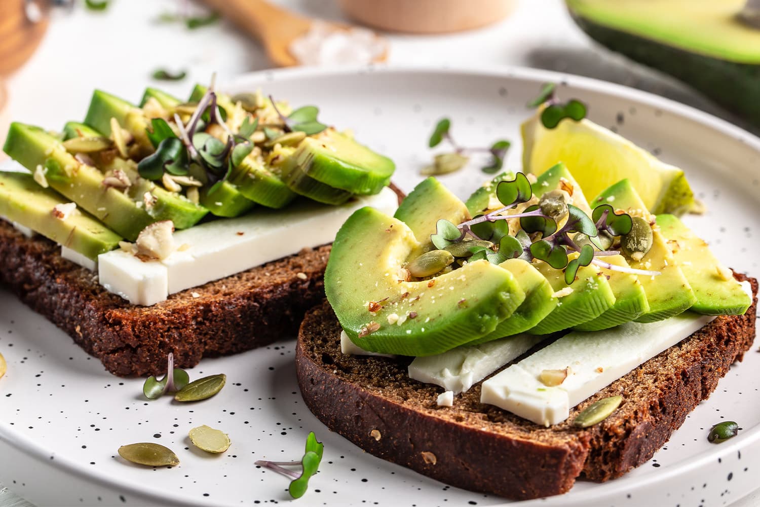 Two slices of dark wholegrain toast topped with sliced avocado, white cheese, microgreens, and pumpkin seeds on a white speckled plate