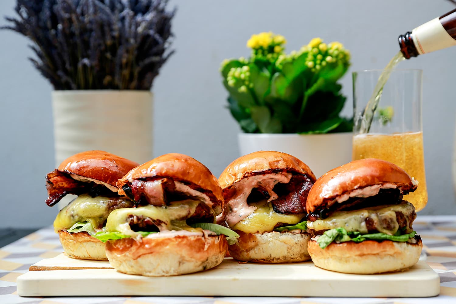 Four artisanal burgers with glazed buns, crispy bacon, and avocado slices arranged on a white board, with potted plants and a glass of beverage in the background.