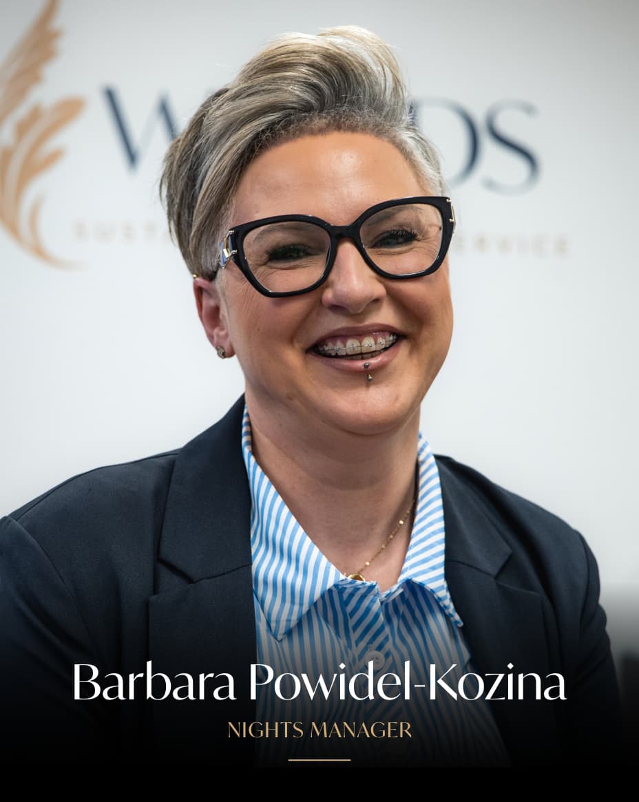 Professional headshot of Barbara Powidel-Kozina, Nights Manager, wearing black-framed glasses, a dark blazer, and blue striped shirt, smiling at the camera.