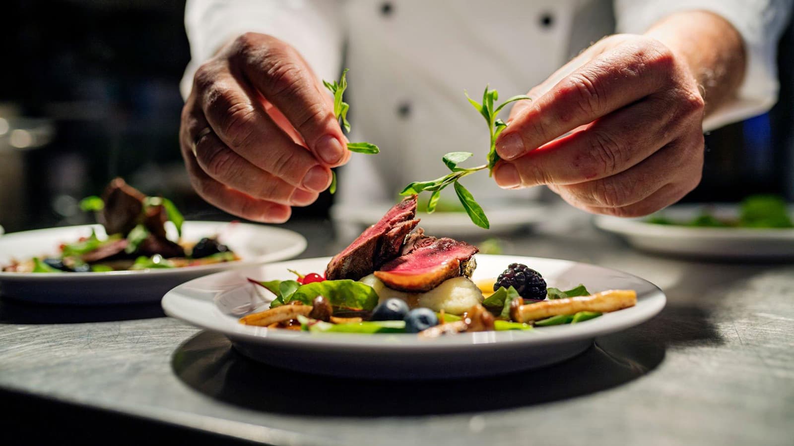 A chef in white chef's coat garnishing a fine dining plate with seared meat, vegetables, berries, and fresh microgreens, with additional plated dishes visible in the background.