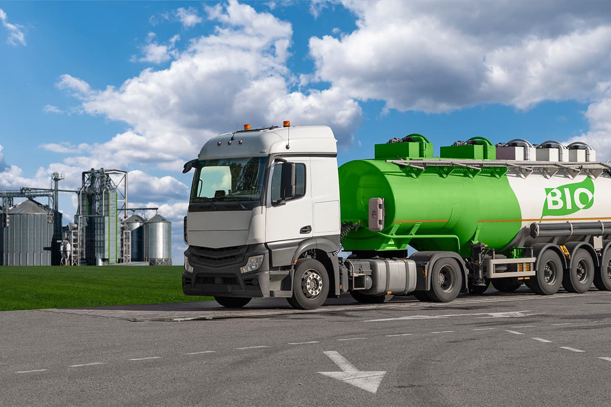 A white and green tanker truck labelled 'BIO' parked at an agricultural facility with grain silos in the background under a blue sky with white clouds.