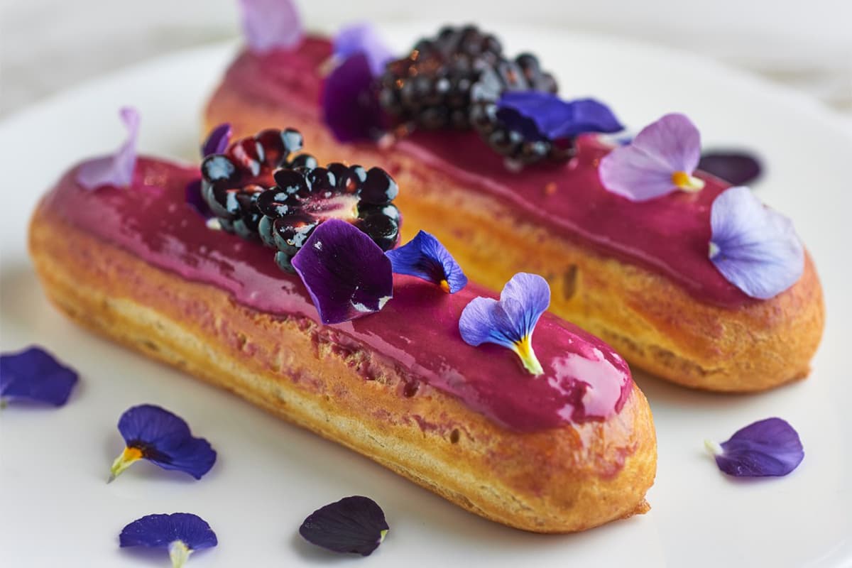 A elegant éclair pastry topped with vibrant magenta glaze, fresh blackberries, and delicate purple and blue edible flowers on a white background.