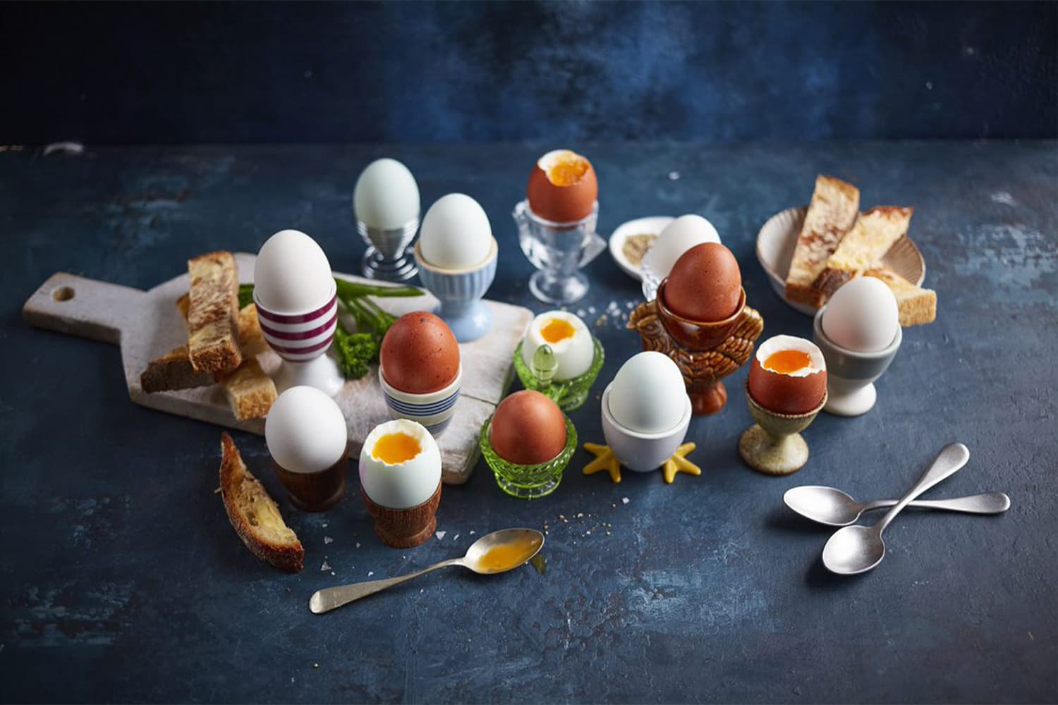 Assorted Clarence Court eggs in decorative egg cups with soldiers, displayed on a dark blue surface with cutlery and fresh parsley garnish.