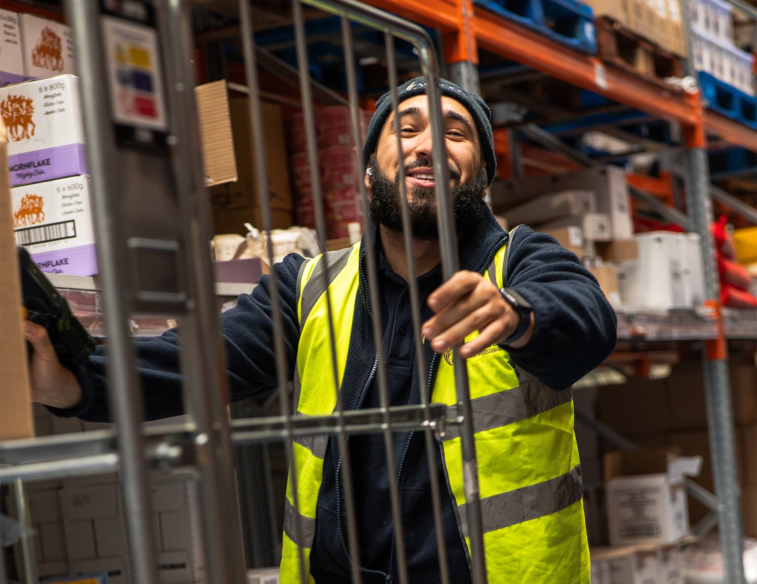 A bearded male warehouse worker wearing a high-visibility yellow and black safety vest and dark beanie hat, smiling and gesturing whilst standing among industrial storage shelves in a distribution centre.