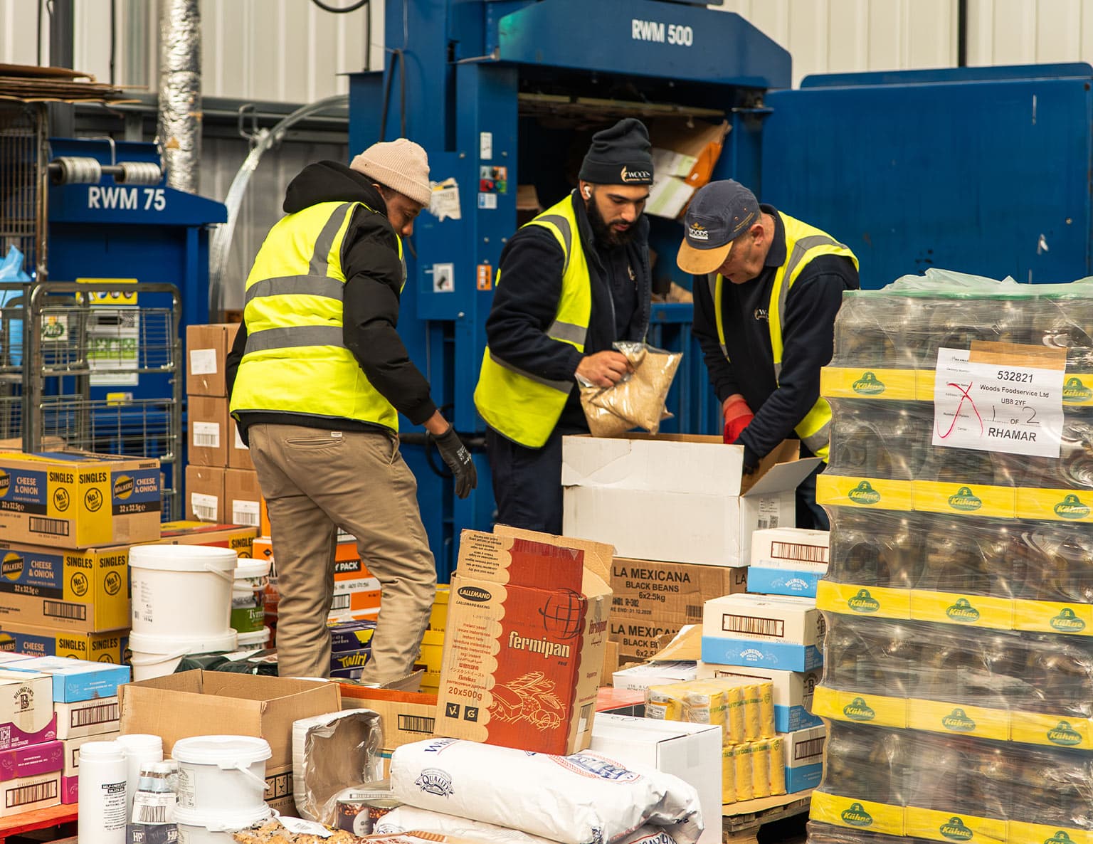 Three workers wearing high-visibility vests sorting and processing food product boxes in an industrial warehouse with blue waste management equipment