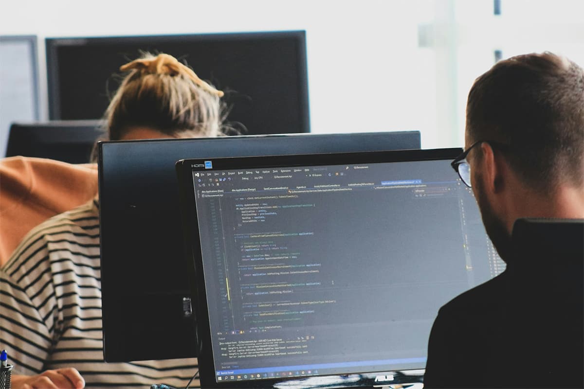 Two software developers working together at a desk, viewing code on a computer monitor in an office environment.