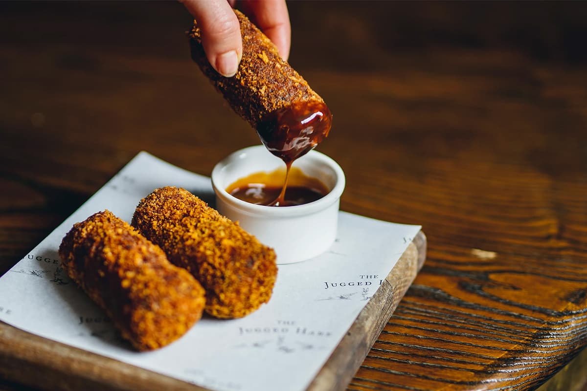 Hand dipping a golden fried croquette into a small white bowl of dark sauce, with two more croquettes on a white plate beside it on a wooden table.