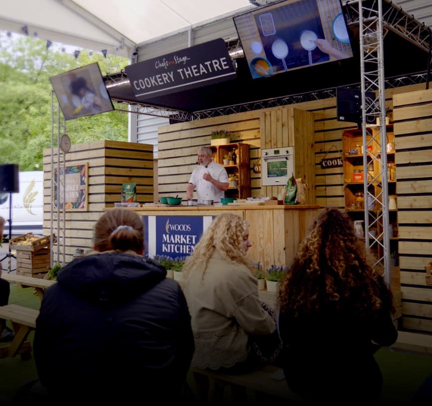 A chef demonstrates cooking techniques on stage at the Cookery Theatre, with three audience members viewing from the foreground. The wooden stage features the Woods Market Kitchen branding and professional lighting rig.