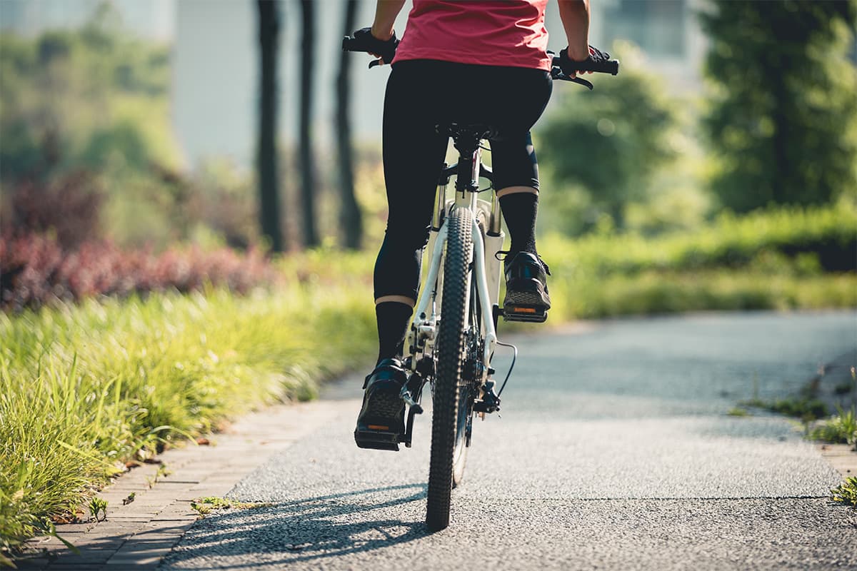 A person in a red and pink top riding a mountain bike on a paved path alongside water and lush green vegetation on a sunny day.