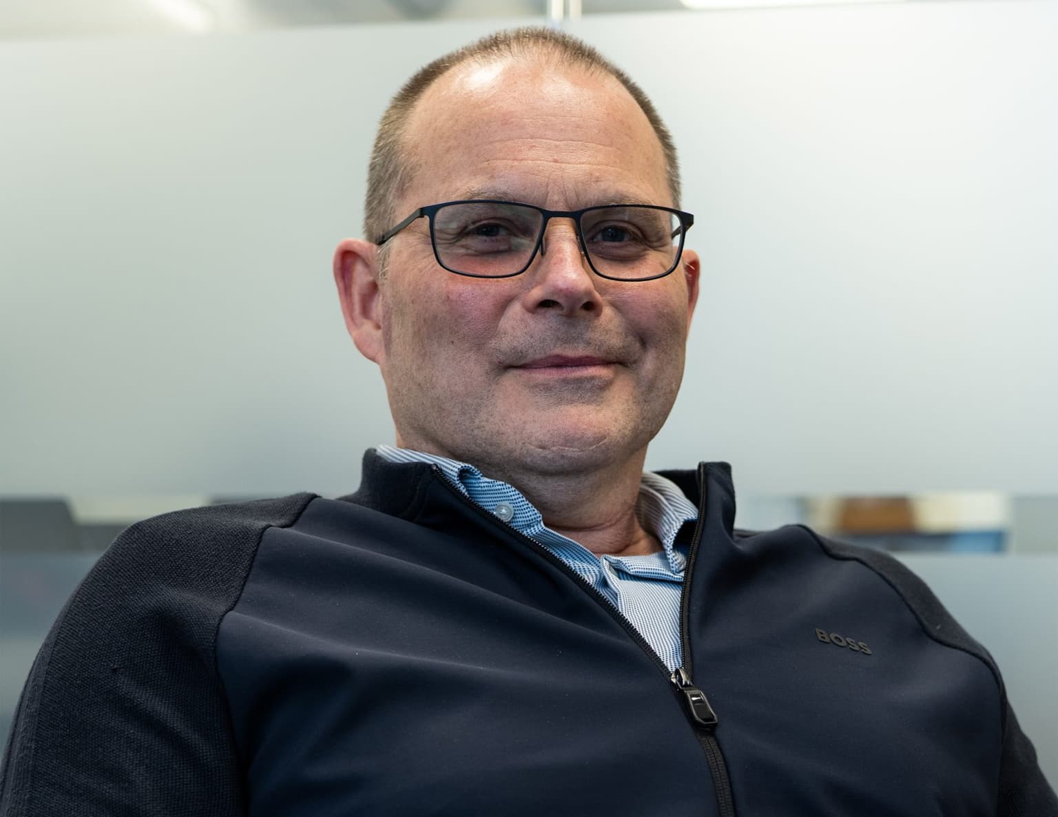 A middle-aged man wearing glasses and a black Hugo Boss jacket over a light blue shirt, smiling at the camera against a plain white background.