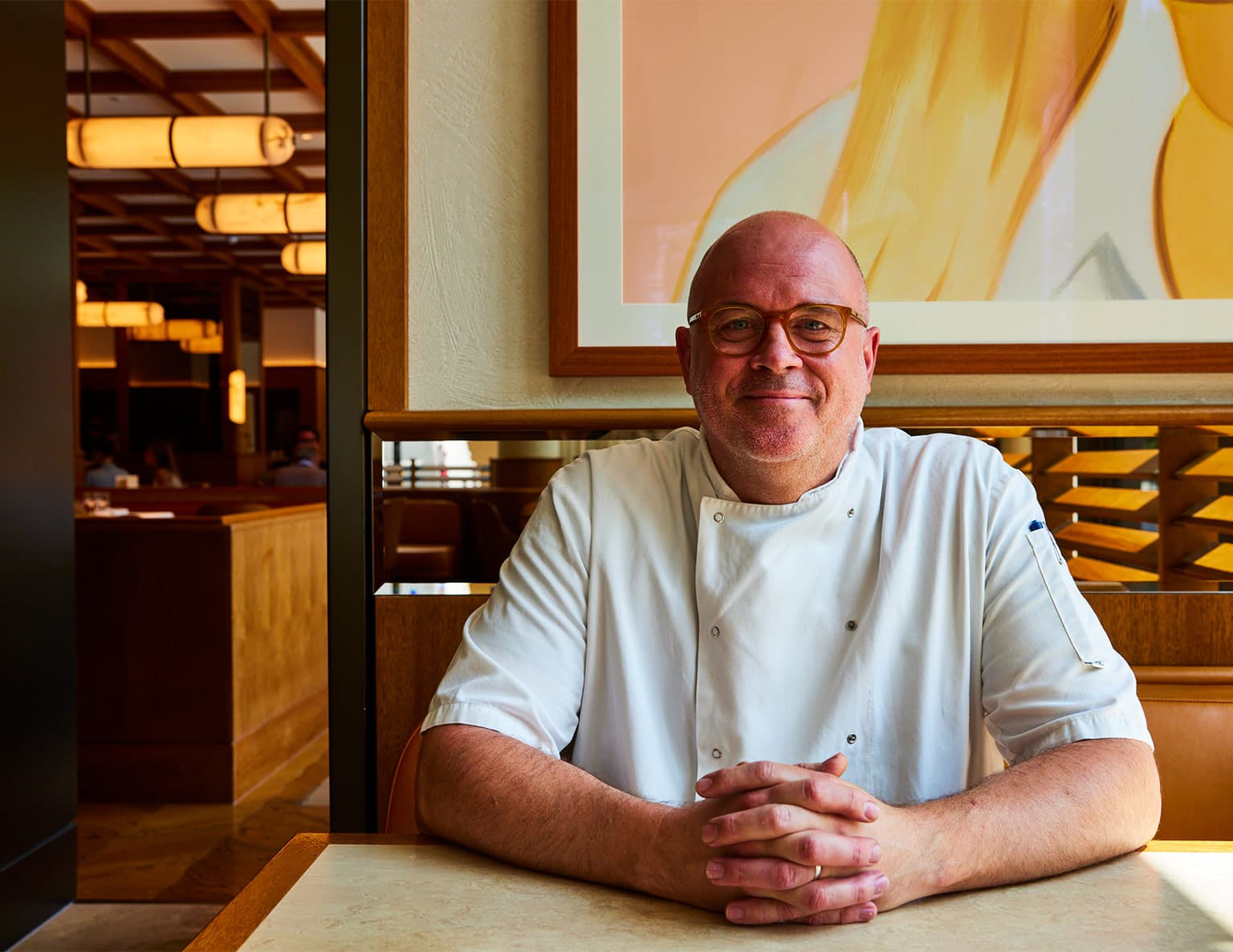 A man wearing glasses and a white chef's coat seated at a table in a modern restaurant interior with warm wooden tones and abstract artwork.