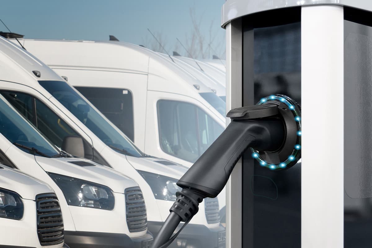 A black and white electric vehicle charging station with blue LED indicators connected to a charging cable, with a row of white commercial vans parked in the background.