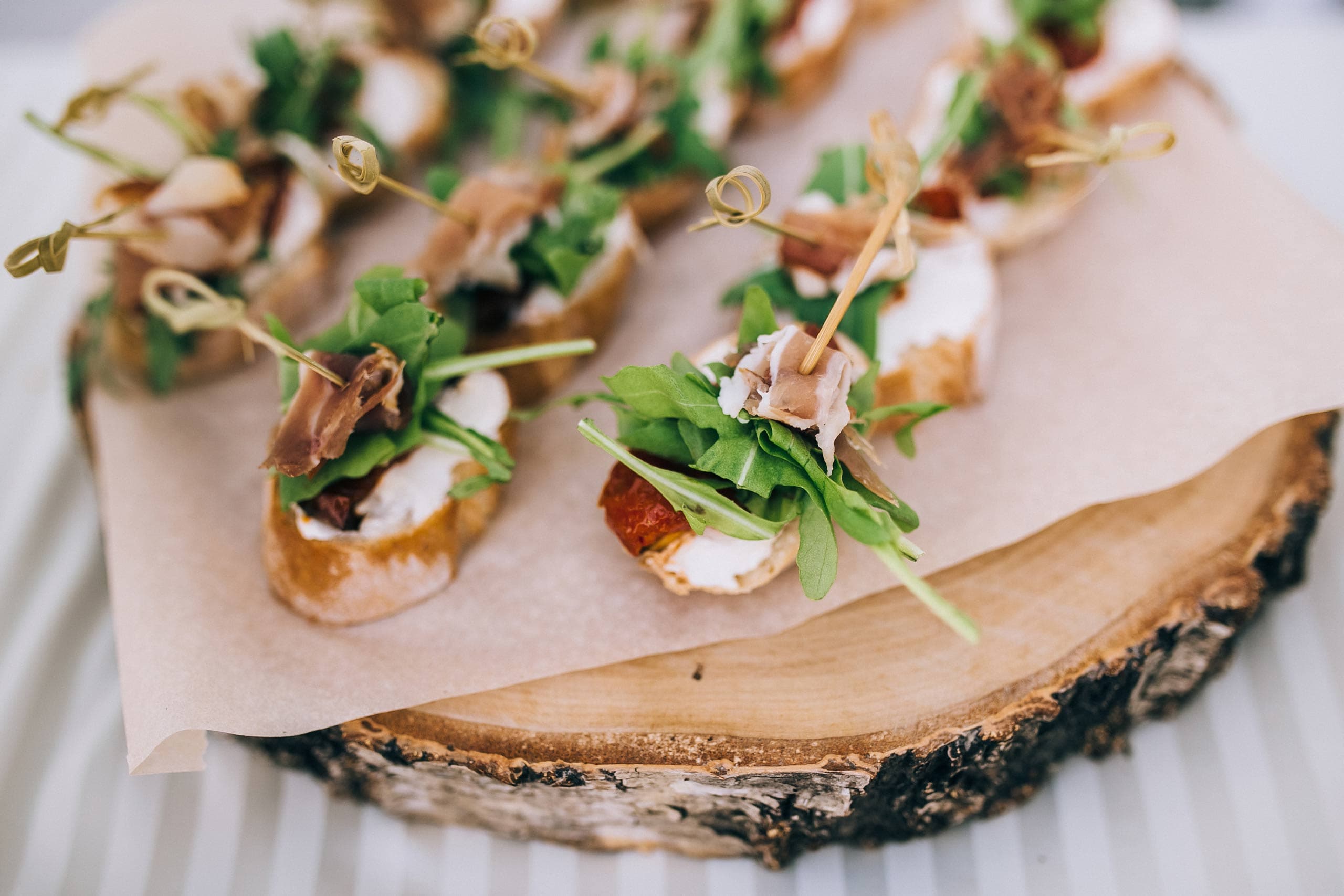 Fine dining canapés arranged on a rustic wooden board with bark edge, each topped with white spread, cured meat, fresh microgreens and decorative picks
