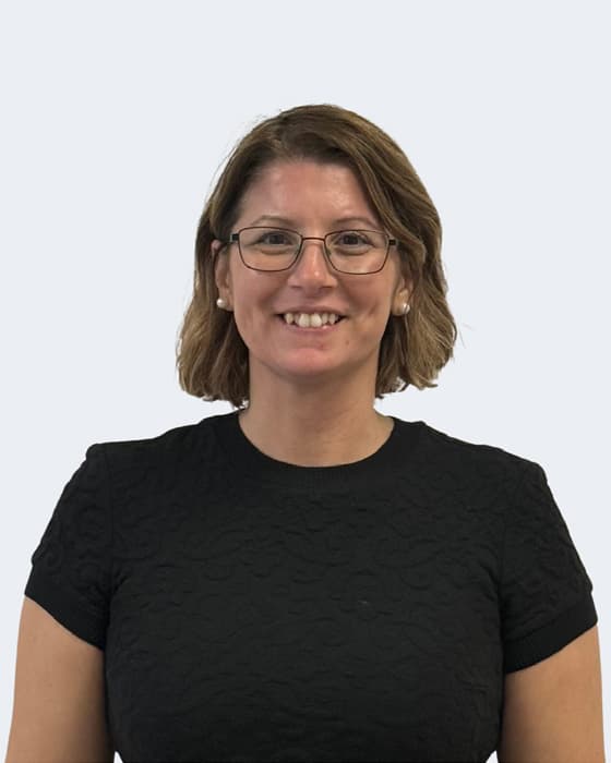 Professional headshot of a smiling woman with shoulder-length brown hair, wearing glasses and a black t-shirt, photographed against a white background.