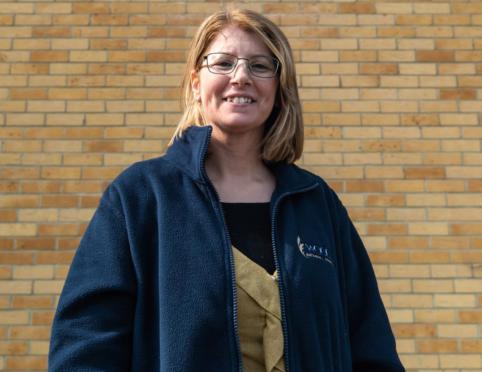 Portrait of a smiling woman wearing glasses and a dark blue fleece jacket over a black and olive-coloured top, photographed against a brick wall background.