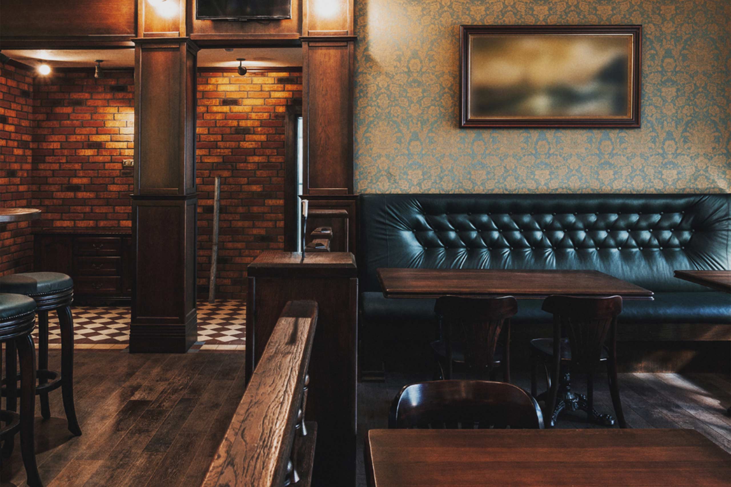 Interior of a contemporary gastropub featuring a black leather button-tufted booth, wooden tables, exposed brick walls, damask patterned wallpaper, and period lighting fixtures.