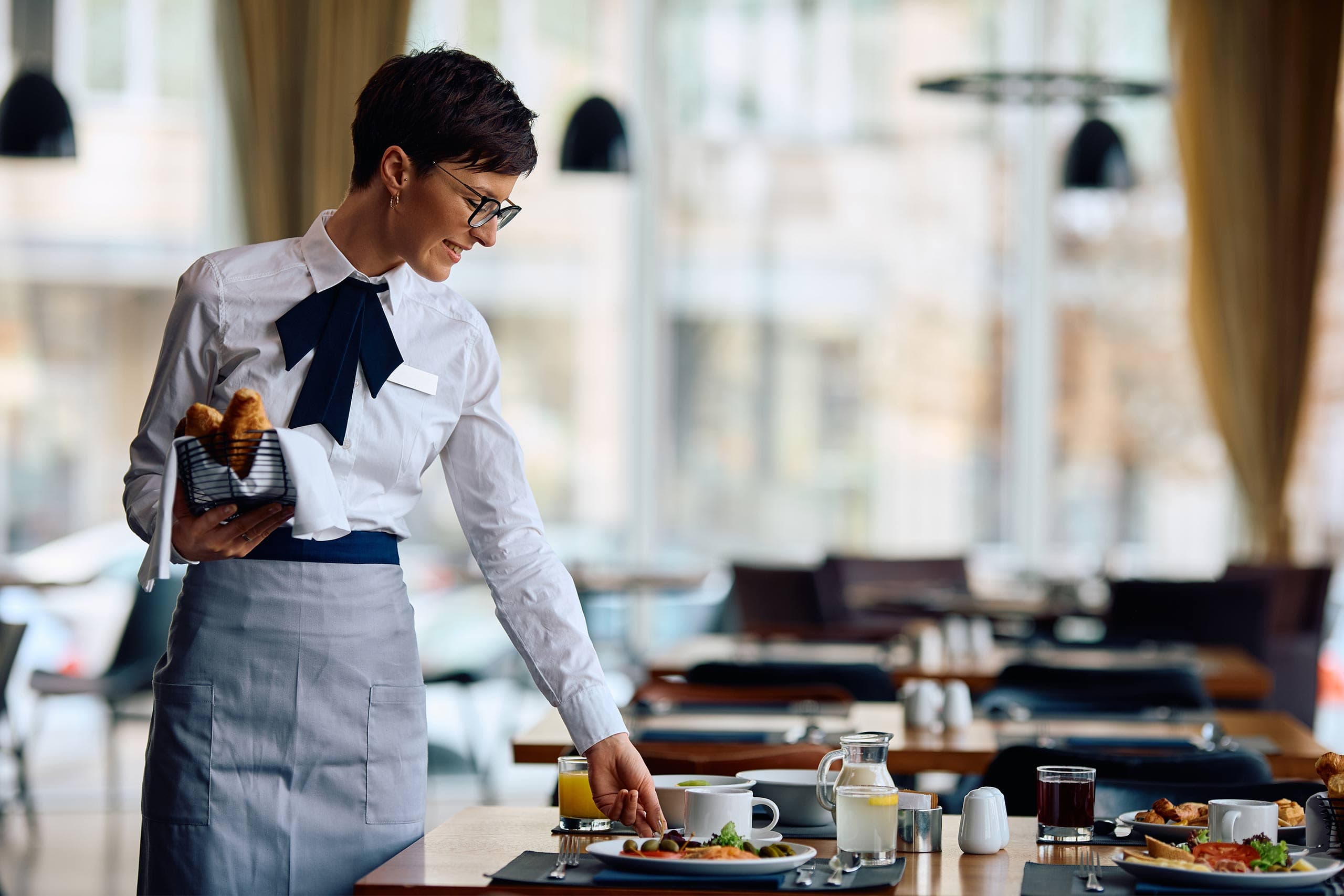 Smiling hotel staff member in formal uniform arranging a breakfast table with pastries, juice, and beverages in an elegant dining room with city views