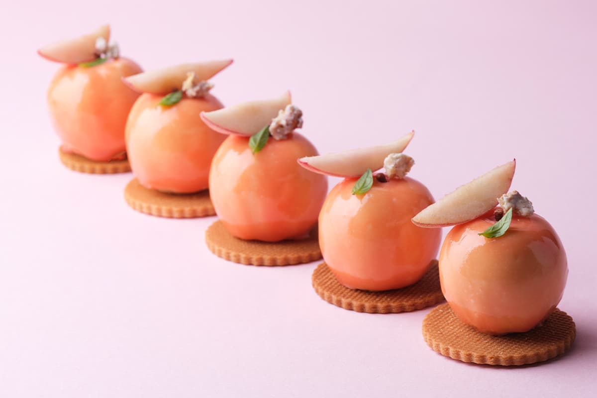 Five apricot-coloured spherical desserts arranged in a diagonal line on a pink background, each topped with white almond petals and a small pink flower garnish, sitting on round biscuit bases.