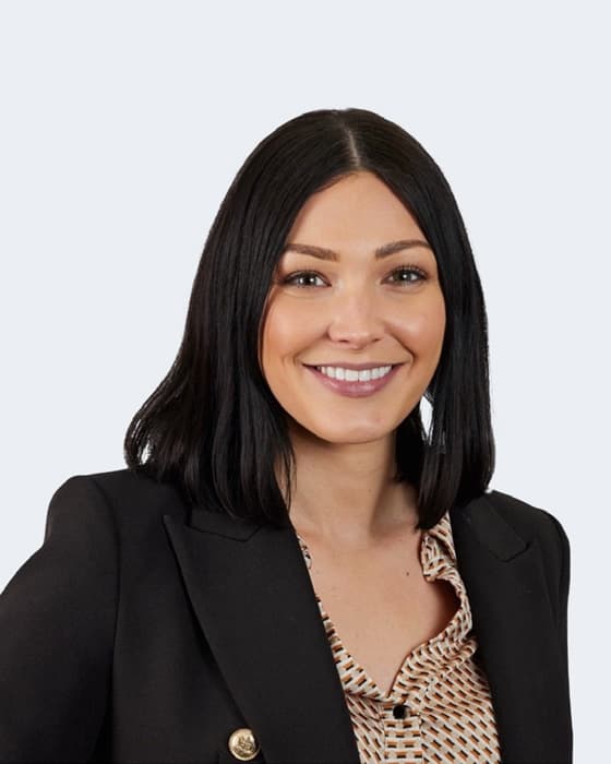 Professional headshot of a woman with long black hair, wearing a black blazer and patterned blouse, smiling against a plain white background.
