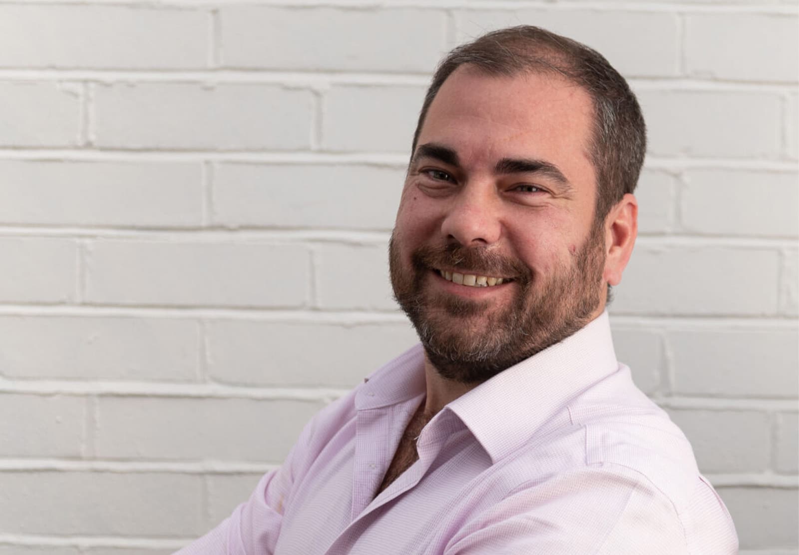 Portrait photograph of a man with a beard wearing a light pink shirt, smiling at the camera against a white brick wall background.