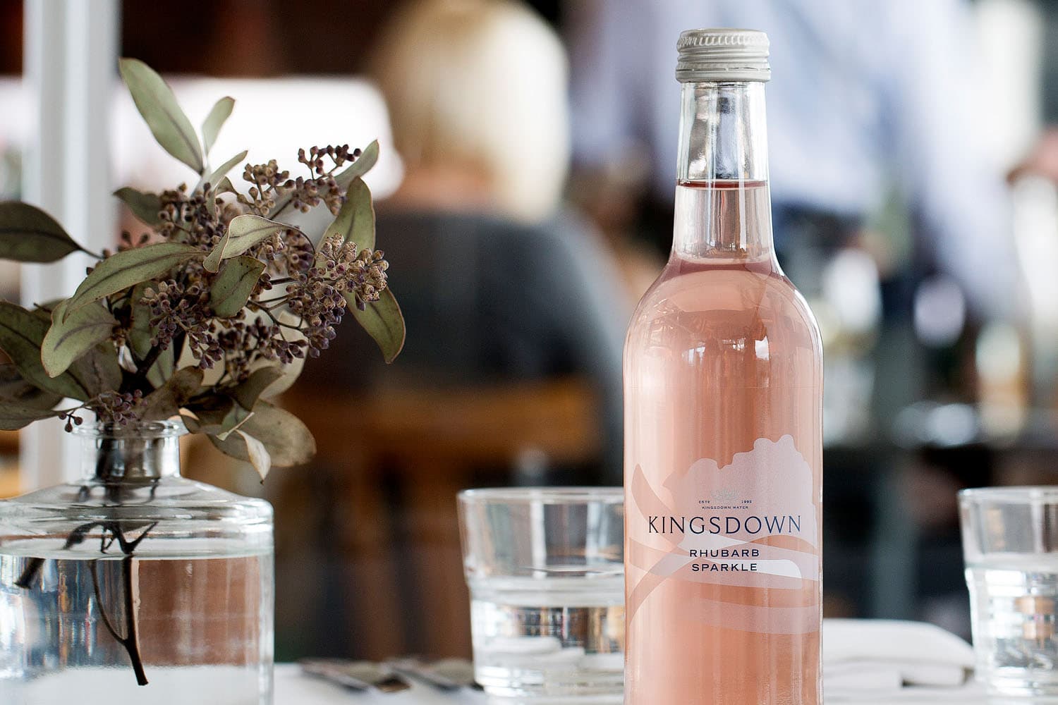 A bottle of Kingsdown Rhubarb Sparkle beverage displayed on a table with a glass vase containing dried flowers and foliage, alongside empty glasses in an elegant table setting.