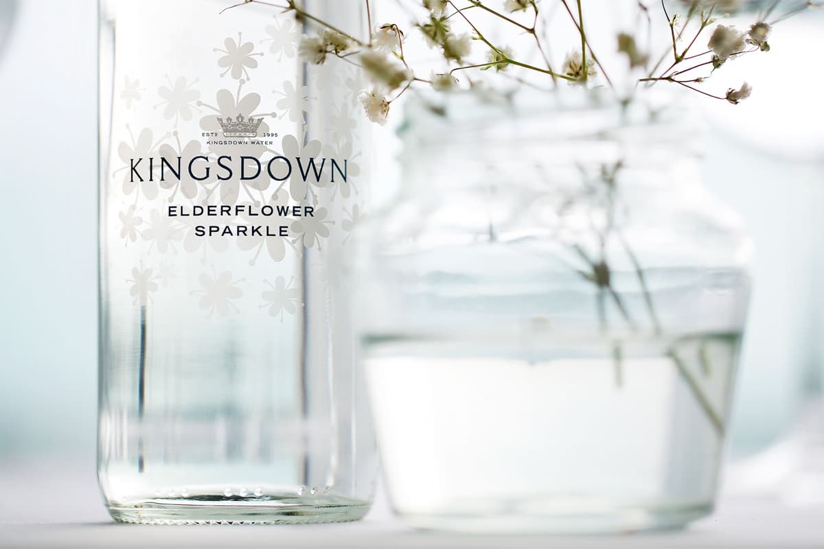 A glass bottle of Kingsdown Elderflower Sparkle water displayed with delicate dried elderflowers in a soft, minimalist setting with natural light.