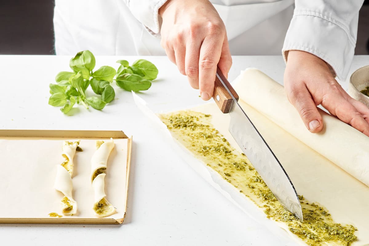 Professional chef in white chef's coat using a knife to slice fresh mozzarella cheese coated with ground pistachios, with fresh basil leaves visible on the work surface.