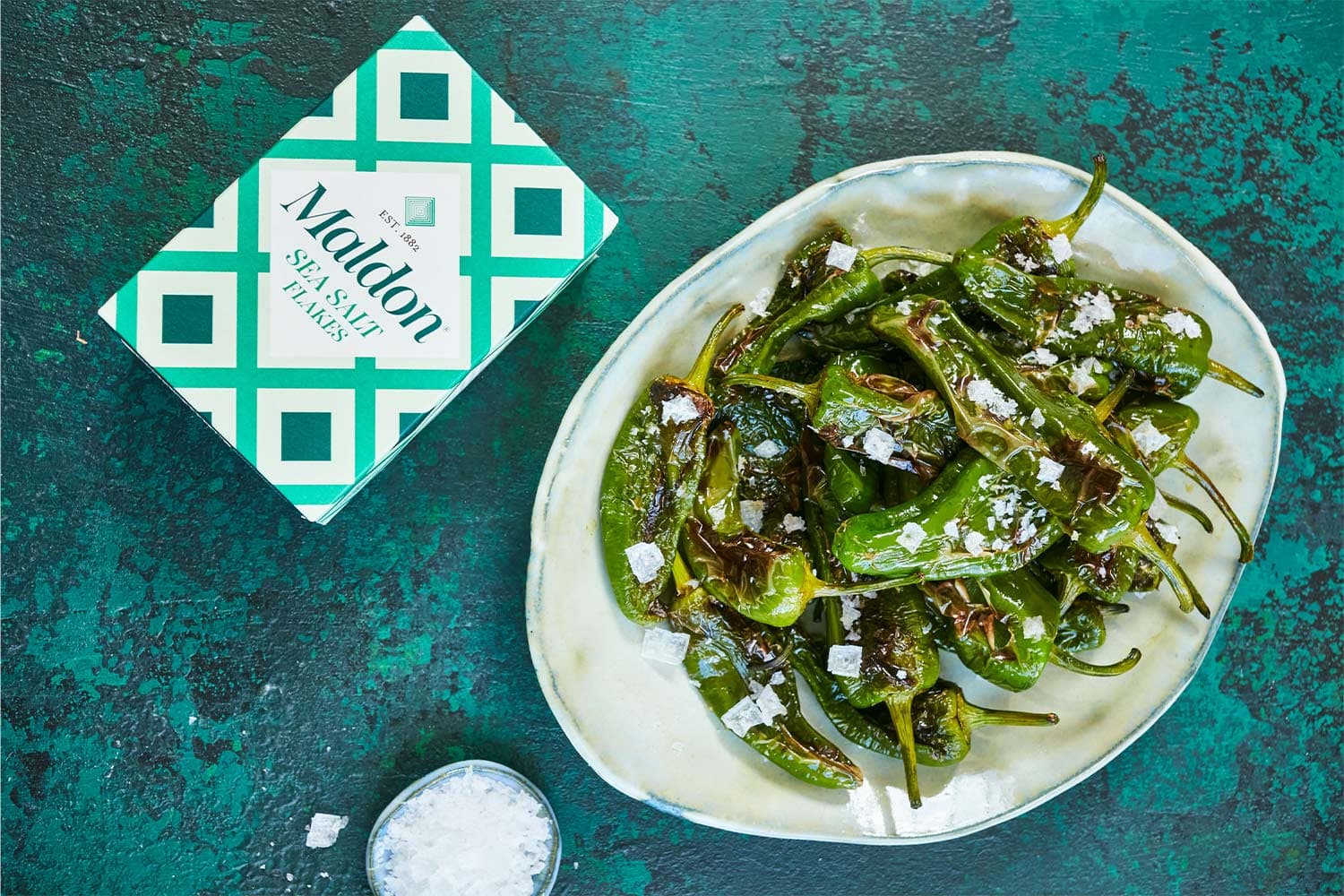 A white bowl containing charred shishito peppers topped with Maldon sea salt flakes, positioned next to a Maldon Salt package on a textured turquoise background.