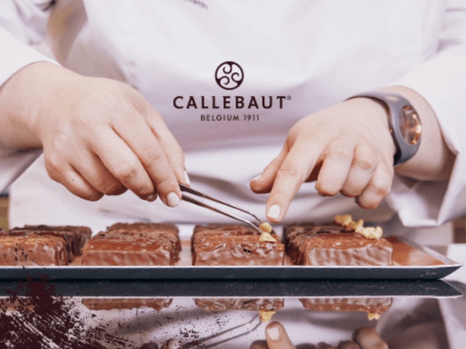 A pastry chef's hands using tweezers to garnish and plate rectangular chocolate pieces with gold leaf, featuring the Callebaut Belgium 1911 logo in the background.