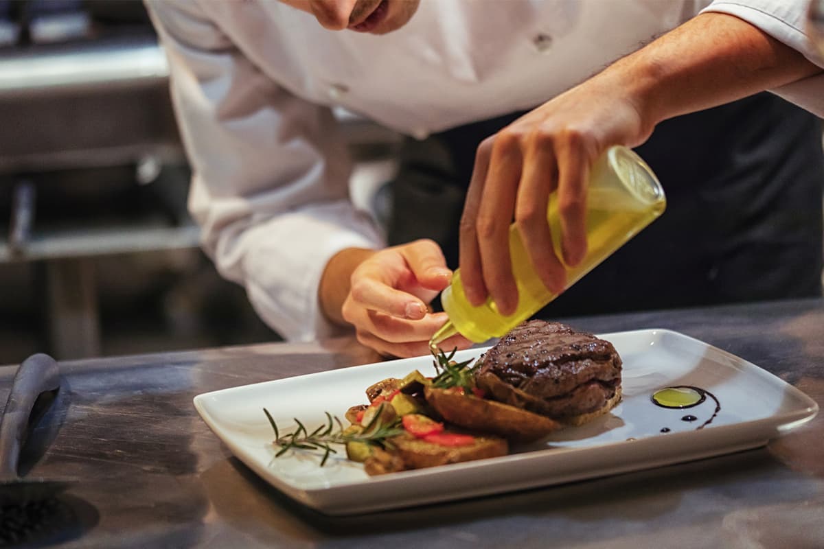 A chef in white chef's coat drizzles golden olive oil from a bottle over a plated steak with roasted vegetables and rosemary garnish.