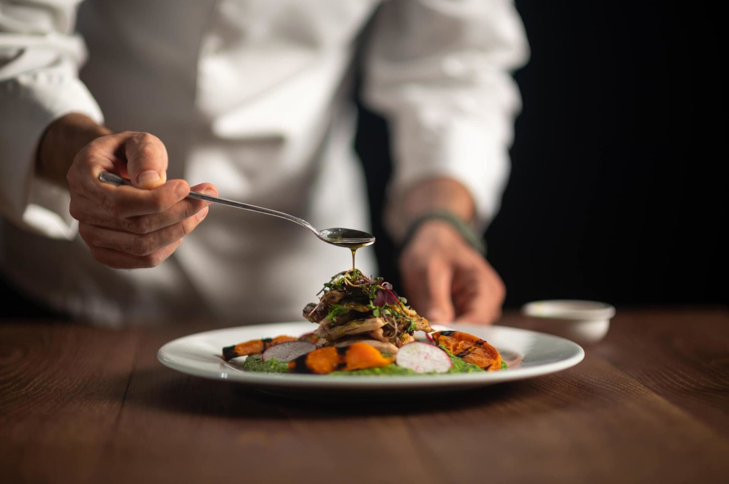 Professional chef in white chef's coat drizzling olive oil over an artfully plated dish featuring roasted vegetables, microgreens, and a white foam element on a white plate.